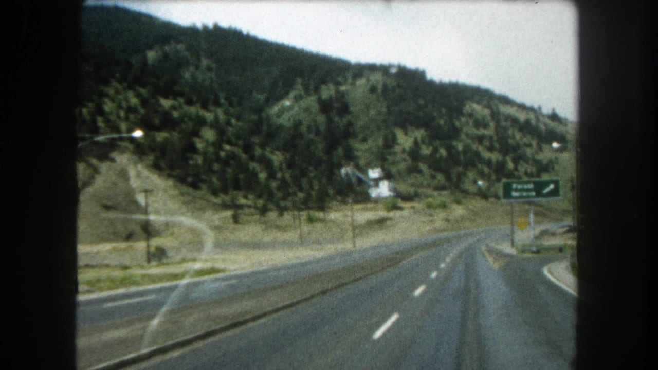 Curving highway through wooded hills with a green roadside sign at right.
