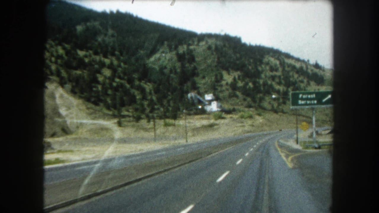 Passenger-view 8mm frame of a Summit County highway curving past wooded mountain slopes, dry roadside ground, and a green Forest Service sign.