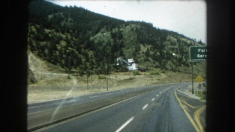1962 Colorado road-travel frame showing the vehicle approaching a curve, with forested slopes, a small white building, and a partially visible Forest Service sign.