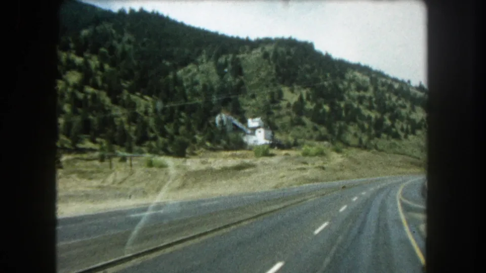 End frame from a moving 8mm car view in Summit County, Colorado, showing a curving highway, wooded hillside, dry verge, and small roadside building.