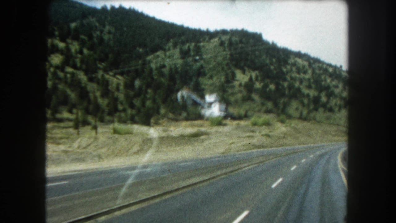 1962 8mm view from a moving car on a Colorado mountain highway, with forested hills, lane markings, glare, and a small white hillside building.