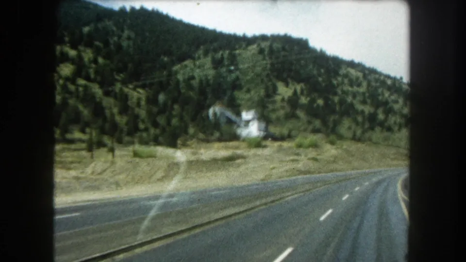 1962 8mm view from a moving car on a Colorado mountain highway, with forested hills, lane markings, glare, and a small white hillside building.