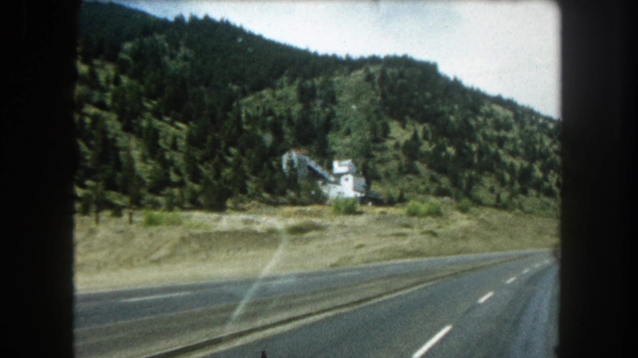 1962 Colorado road footage shows a highway bending through pine-covered hills, with a white mountainside structure and windshield reflections.