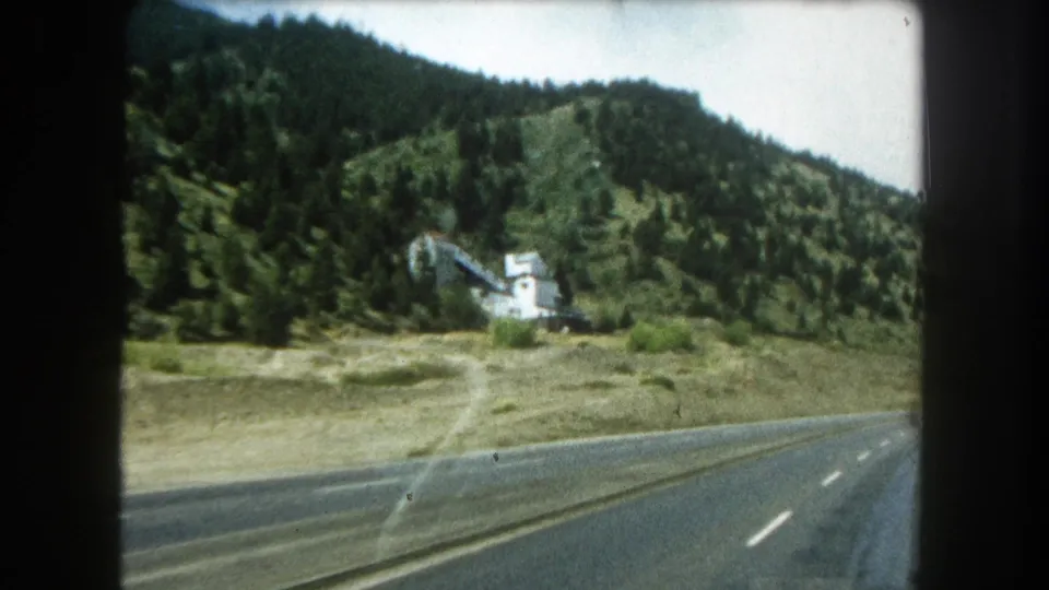 1962 8mm frame from inside a moving car, showing a Colorado mountain highway, pine slopes, and a small white building near the road.
