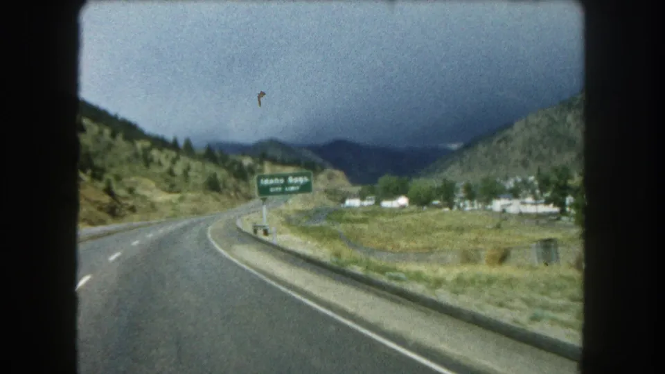 1962 Colorado highway footage shows a green Idaho Springs exit sign, distant mountains, roadside buildings, and dark clouds ahead.