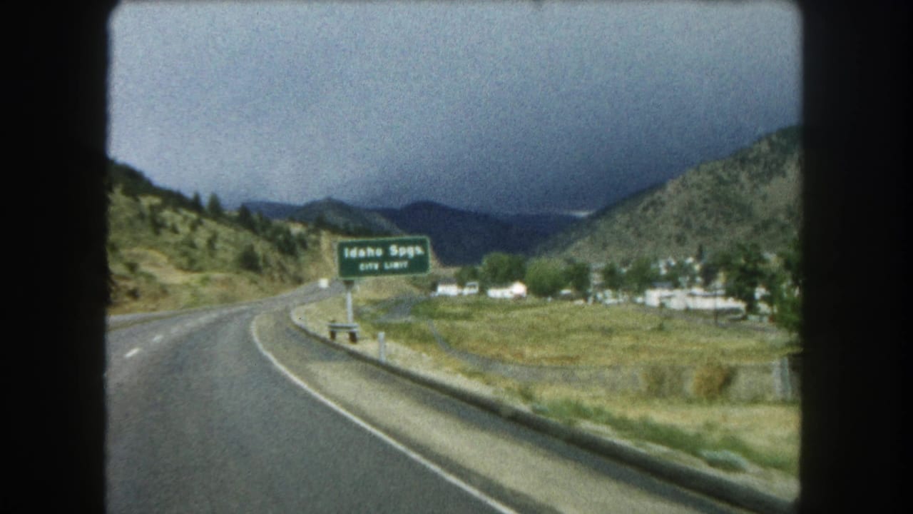 Curving highway beside an Idaho Springs city-limit sign with hills and cloudy mountains beyond.