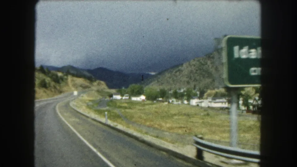 The Idaho Springs sign moves past the camera, with roadway, guardrail, open land, and hills still visible.