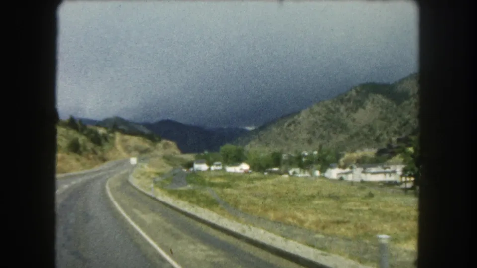 After the sign, the moving-vehicle view shows the Colorado highway corridor, roadside buildings, hills, and clouded mountains.