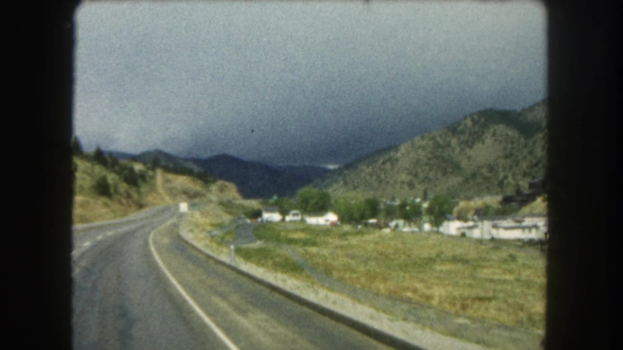 Curving mountain highway beside a grassy valley under dark storm clouds.