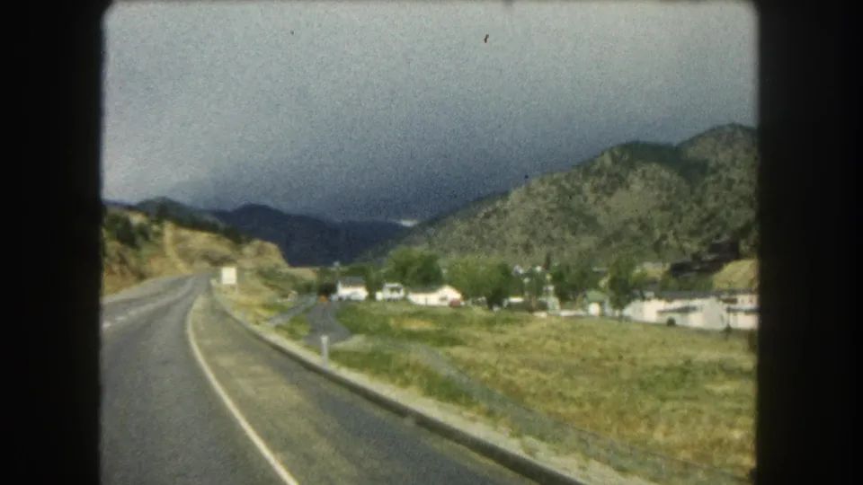 A 1962 8mm travel frame shows a Colorado mountain road, open valley ground, distant homes, and dark clouds over the ridgeline.