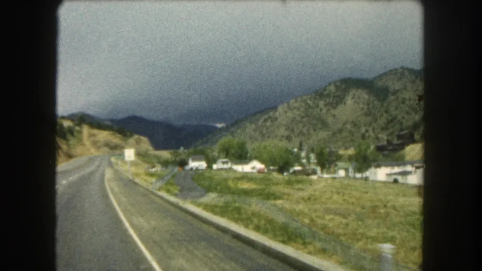 Summit County, Colorado road-view frame from 1962, with a valley settlement, green trees, mountain slopes, and storm-darkened sky.