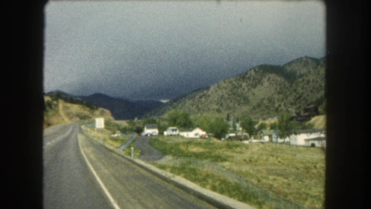 1962 Summit County road view with a paved highway, roadside grass, small houses, and forested Colorado hills in the distance.