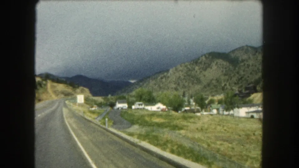 1962 Summit County road view with a paved highway, roadside grass, small houses, and forested Colorado hills in the distance.