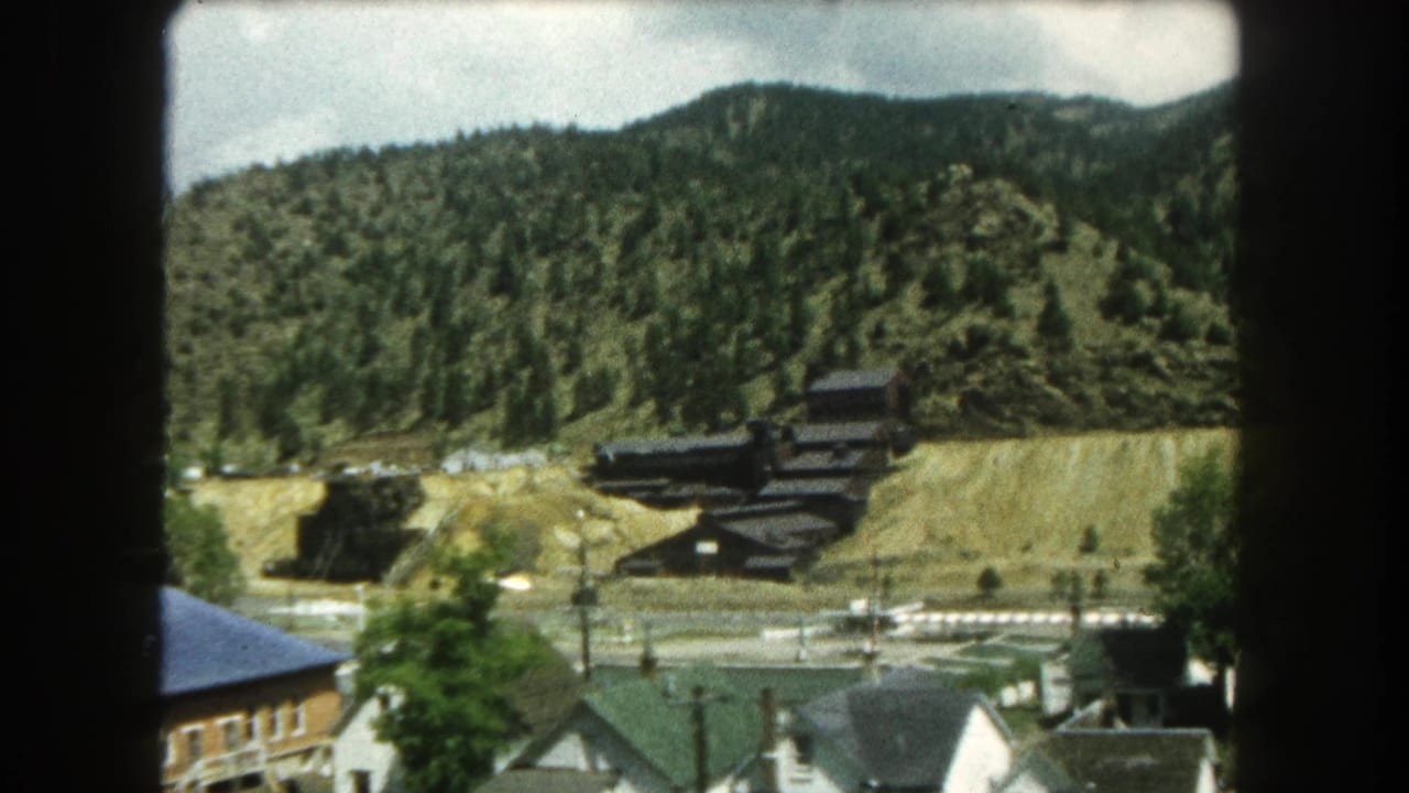 1962 8mm view in Summit County, Colorado showing town rooftops, roads, and a dark hillside structure beneath forested mountains.