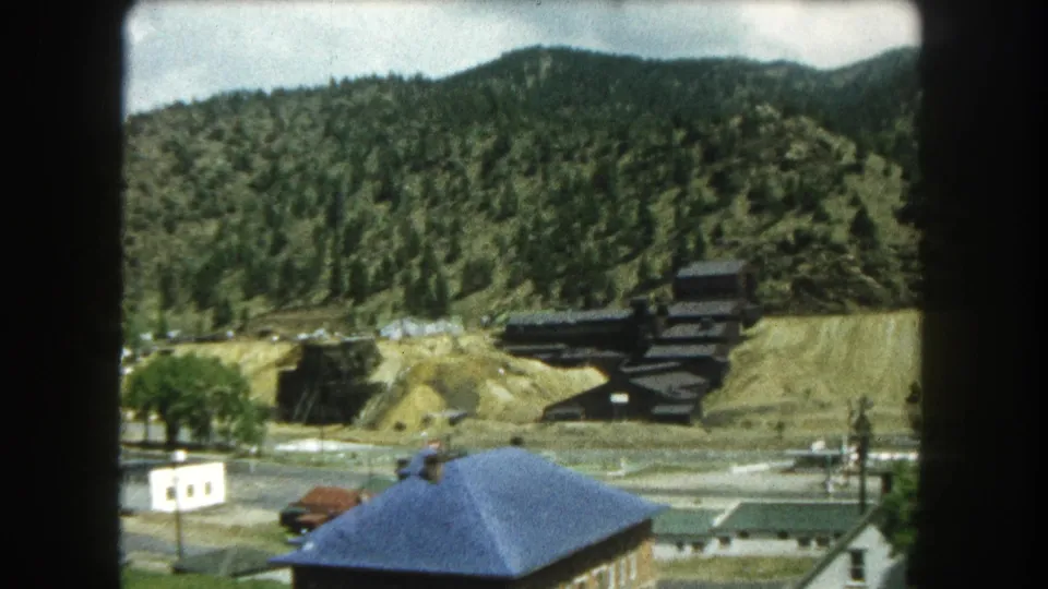 1962 home-movie frame of Summit County rooftops, trees, roads, and a dark hillside structure against mountain forest.