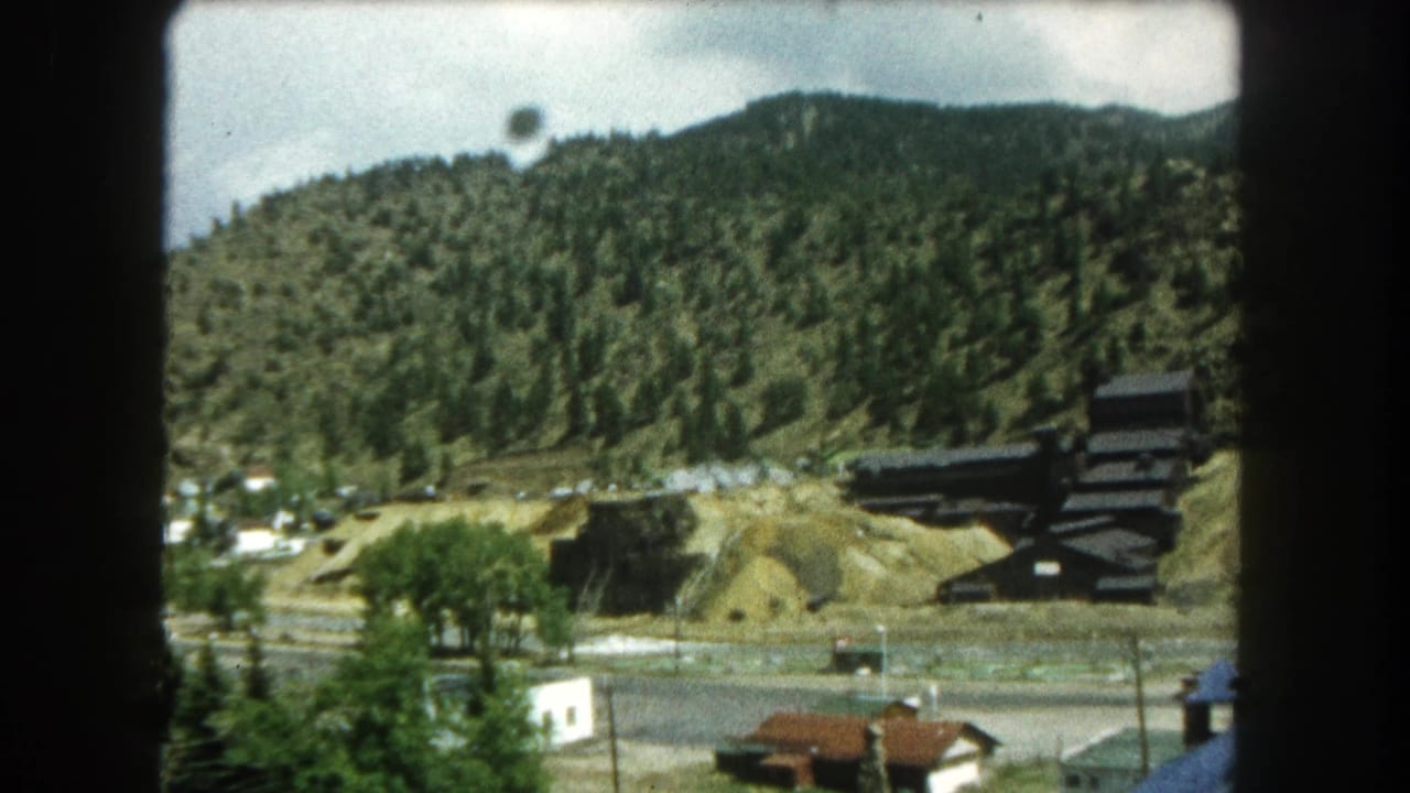 1962 Summit County, Colorado home-movie frame showing houses, a road, trees, and a dark industrial structure on a wooded hillside.