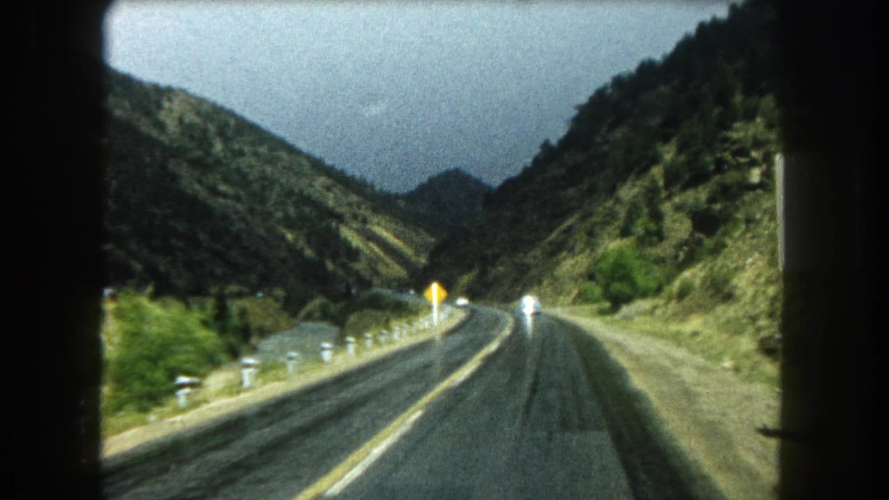 1962 Summit County, Colorado 8mm view from inside a car looking down a wet two-lane mountain road between rocky slopes.