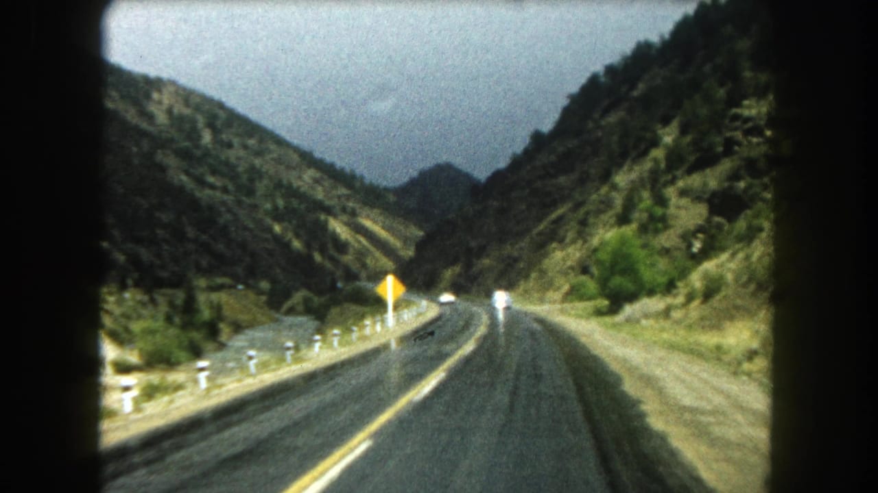 Passenger-view 8mm frame of a wet Summit County mountain road, yellow roadside sign, and oncoming traffic under an overcast sky.
