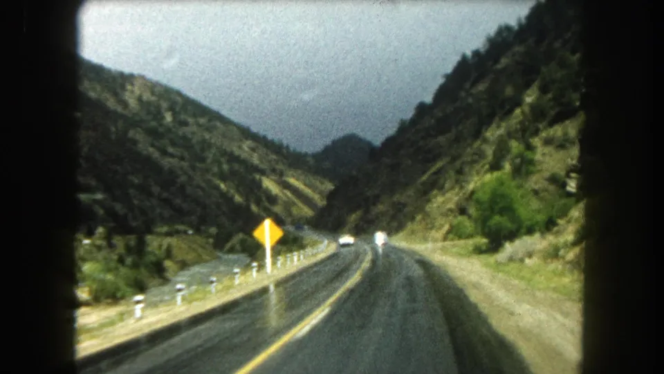 1962 Colorado home-movie frame showing a wet two-lane mountain road, roadside sign, guard posts, and valley slopes from a moving car.