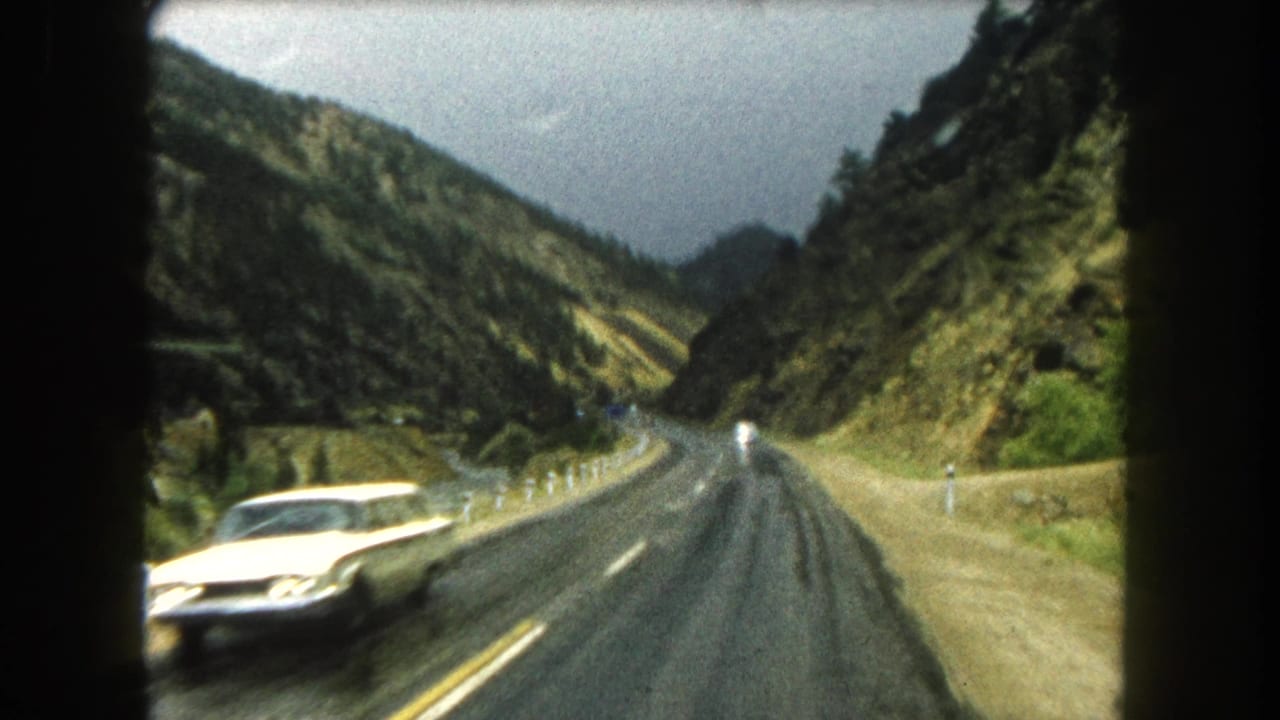 Oncoming white car passes on a wet two-lane road between green and rocky mountain slopes.