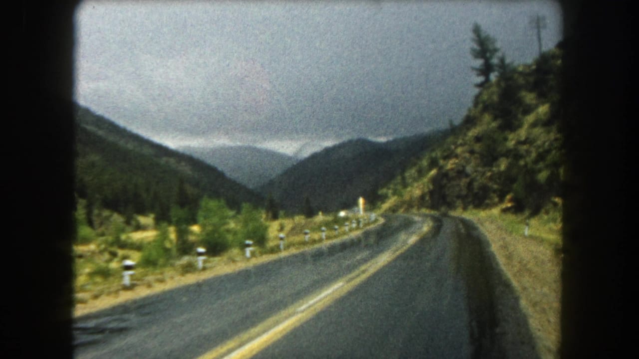 1962 8mm home-movie view of a wet two-lane road through Summit County mountain terrain with evergreen slopes ahead.
