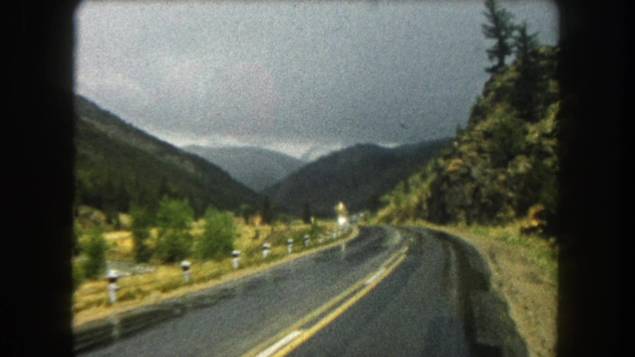 Passenger-view 8mm frame of a Summit County mountain road bending past forest and rocky roadside terrain in 1962.