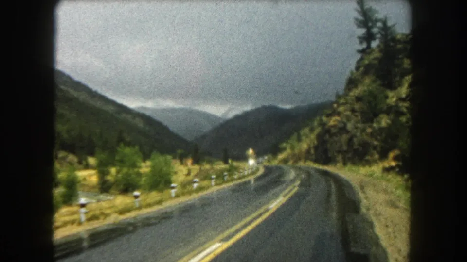 Passenger-view 8mm frame of a Summit County mountain road bending past forest and rocky roadside terrain in 1962.