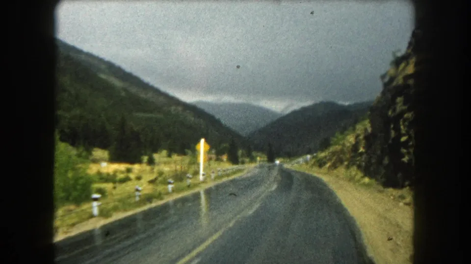 A distant white car travels along a wet Summit County mountain road in this 1962 8mm passenger-view frame.