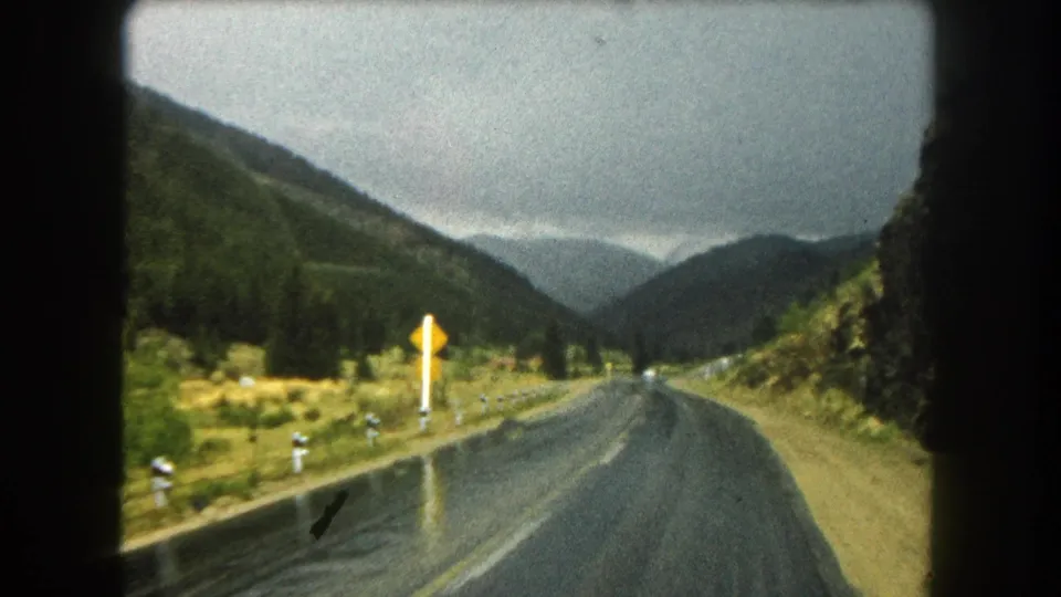 1962 8mm view from a moving vehicle on a wet Summit County, Colorado mountain road, with forested slopes, roadside posts, and a yellow warning sign ahead.