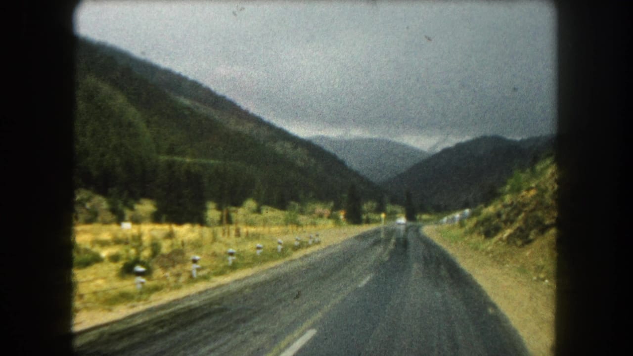 Forward-facing 1962 8mm travel view along a damp Summit County mountain road, with a valley opening between tree-covered slopes and faint vehicles in the distance.