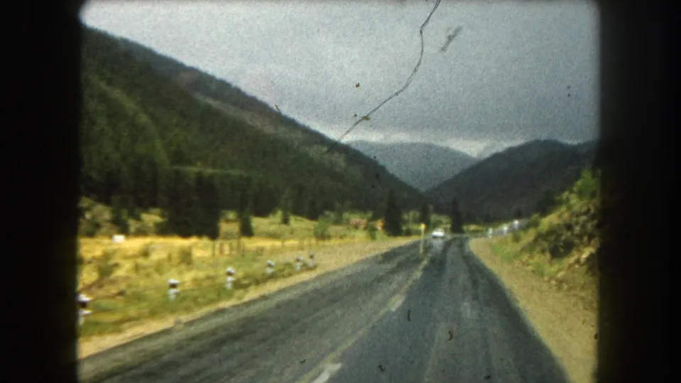 1962 home-movie frame from a moving car on a wet Summit County, Colorado road, showing roadside markers, dark pavement, forested slopes, and distant traffic under heavy clouds.