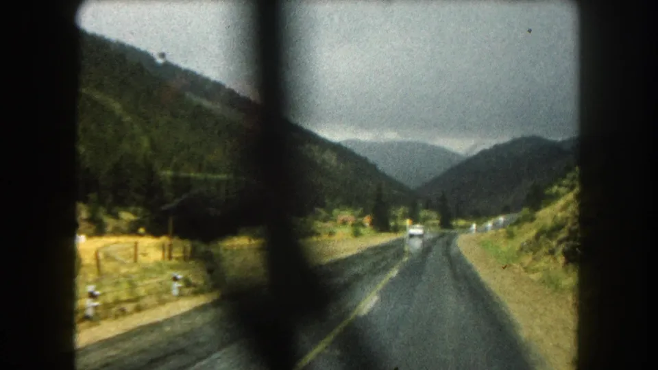 Late-frame 1962 8mm view along a Summit County mountain highway, with wet pavement, forested slopes, distant vehicles, and visible gate or frame shadowing across the scan.