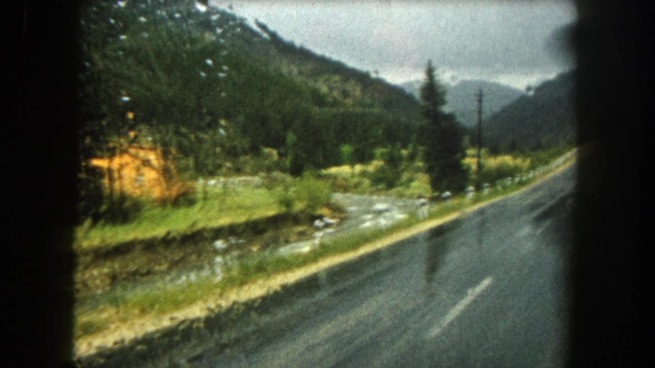 Passenger-view 8mm frame from Summit County showing wet pavement, a stream, evergreen trees, and distant mountains in rainy weather.
