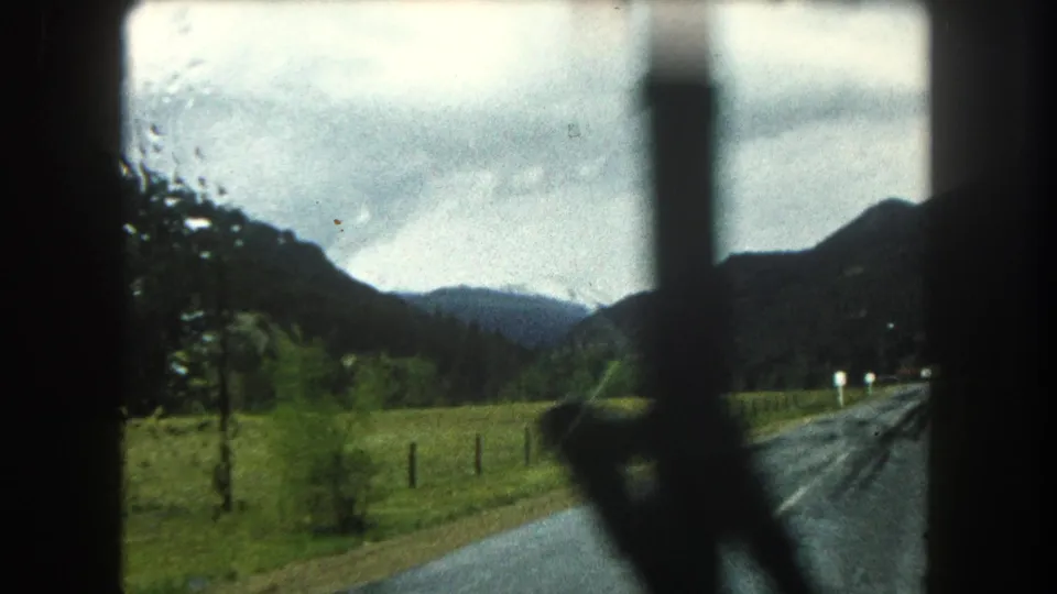 Rainy 1962 Colorado travel view with a blurred windshield wiper or window post over fields, roadside markers, and dark mountain slopes.