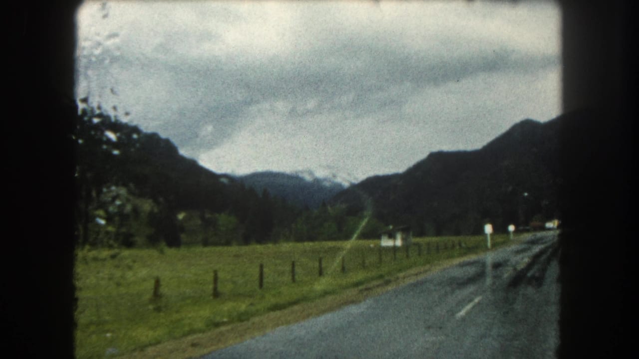 1962 Summit County, Colorado home-movie view of a wet road, pasture, fence posts, and mountains under heavy clouds.