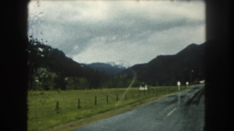 1962 Summit County, Colorado home-movie view of a wet road, pasture, fence posts, and mountains under heavy clouds.