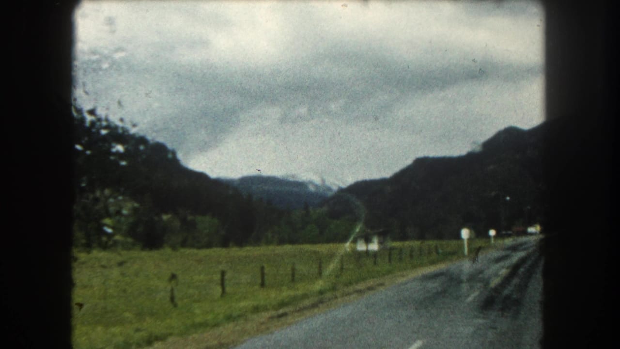 Moving-car view of a wet Summit County road with green fields, roadside markers, forested slopes, and distant mountains.