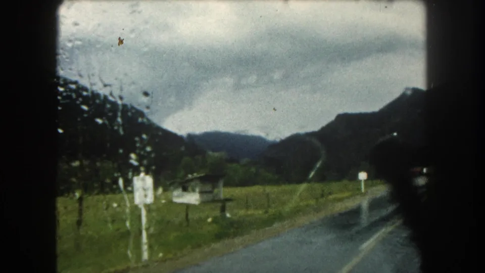 Rain-speckled 8mm frame of a wet mountain road, roadside signs, open field, and dark hills in Summit County, Colorado.