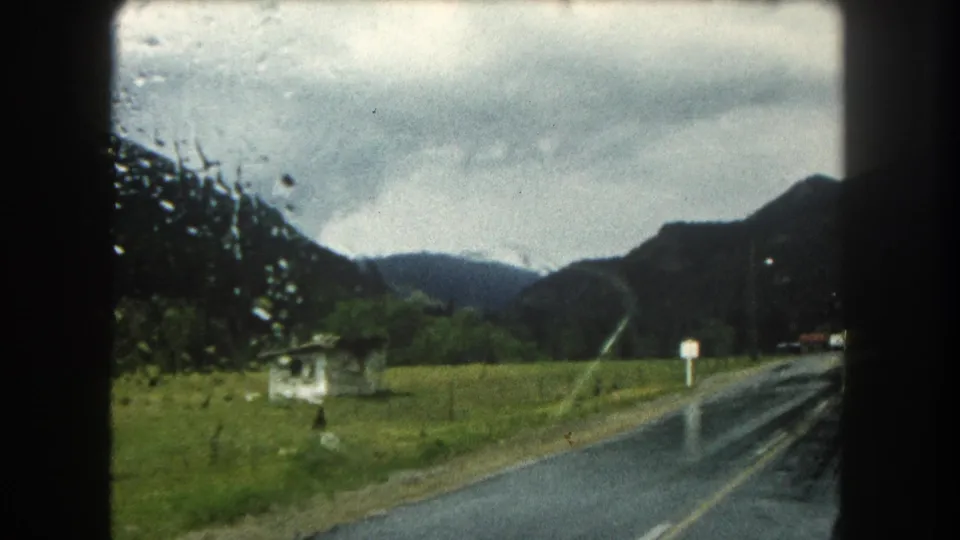 Final frame of the rainy 1962 mountain drive, showing a wet road, pasture, roadside posts, and cloud-covered peaks.