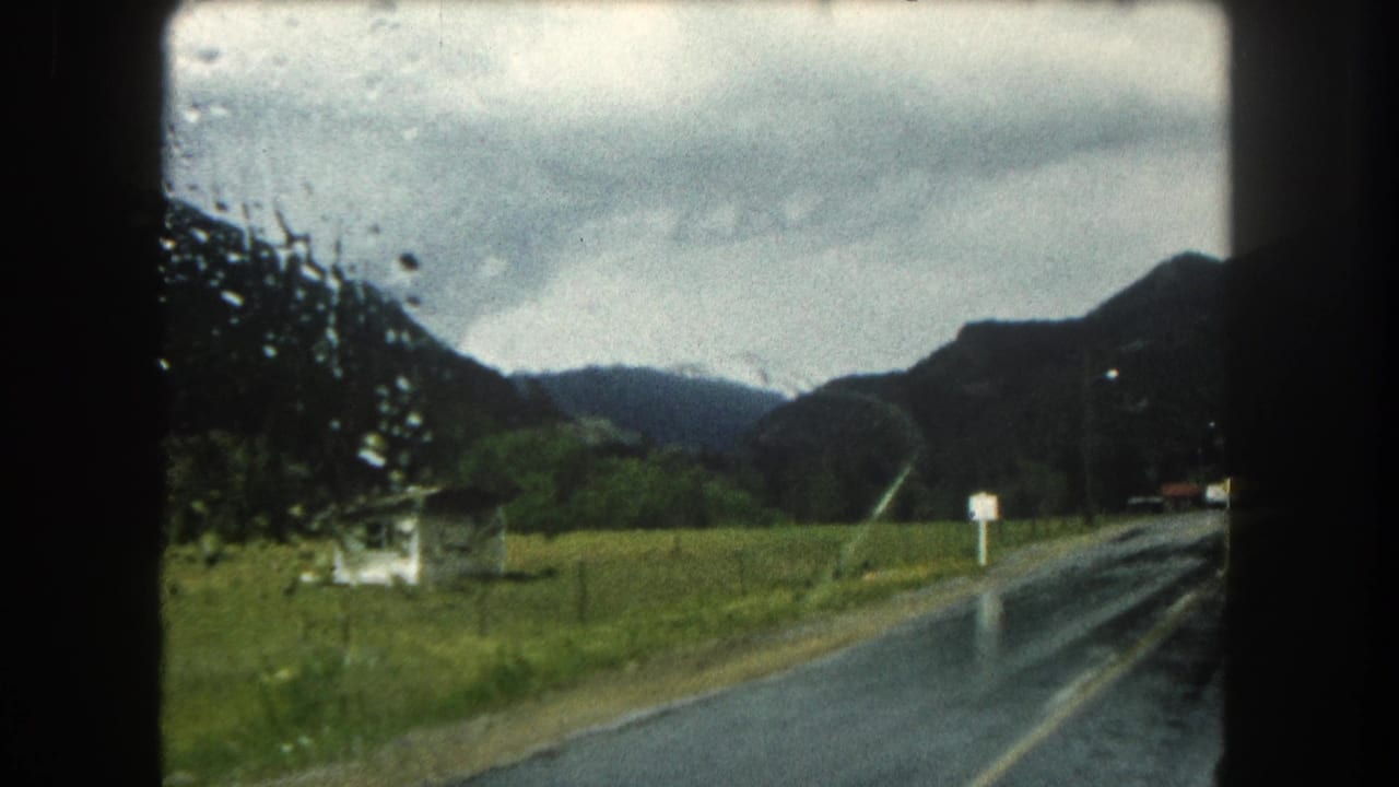 Rainy two-lane road beside green pasture with mountains under an overcast sky.