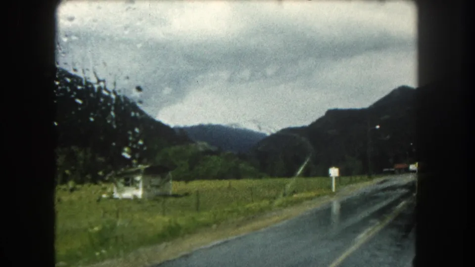 1962 Summit County, Colorado home-movie frame showing a wet rural road, pasture, signs, and distant mountains through rain-speckled glass.