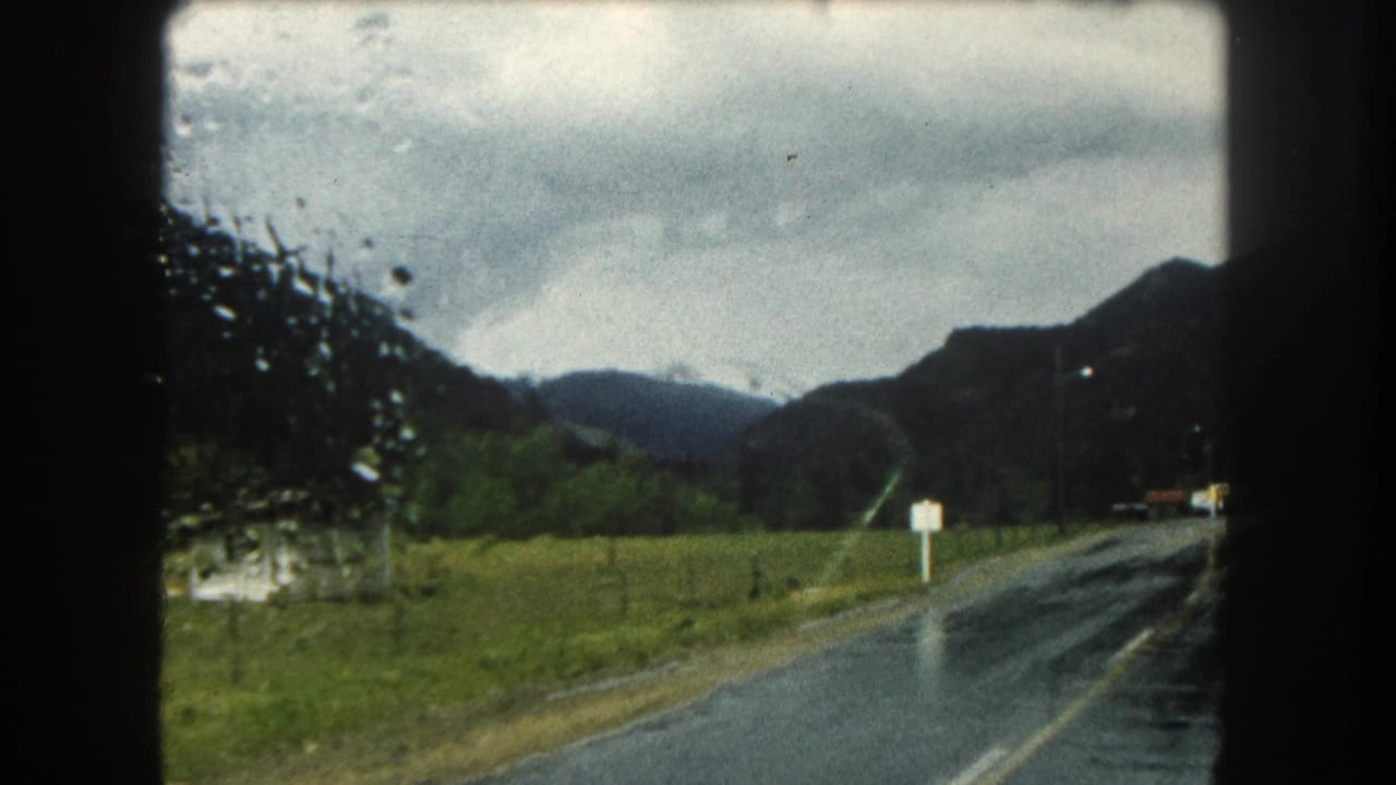 Wet mountain road with pasture, roadside signs, and dark hills in the distance.
