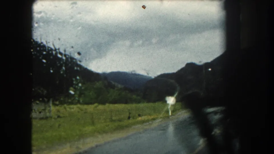 1962 Summit County, Colorado frame with motion blur from a moving vehicle, showing a wet road, pasture, sign, and mountain background.
