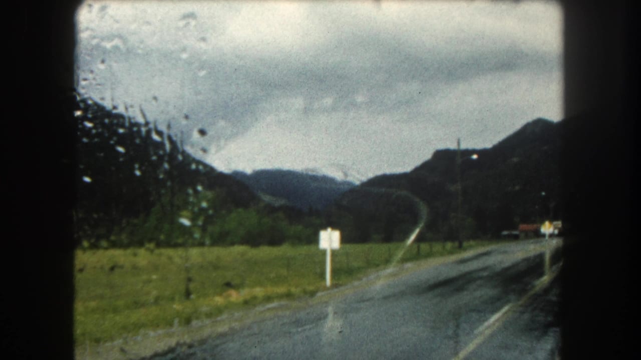 1962 Summit County, Colorado road view through moisture, with a two-lane highway, roadside signs, green slopes, and cloudy mountains.