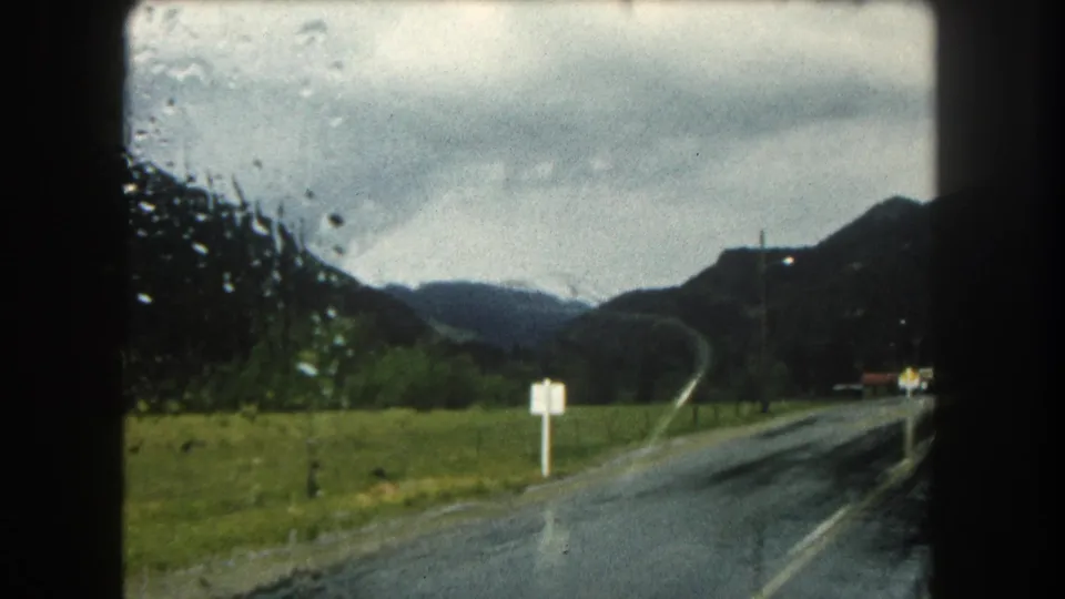 1962 Summit County, Colorado road view through moisture, with a two-lane highway, roadside signs, green slopes, and cloudy mountains.
