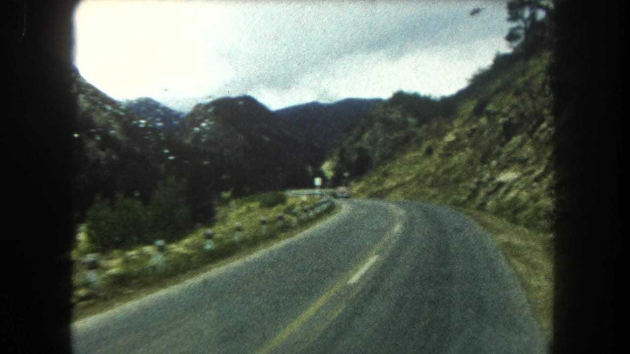 A 1962 Colorado mountain highway curves through green terrain, with guardrail posts, forested slopes, and overcast sky visible ahead.