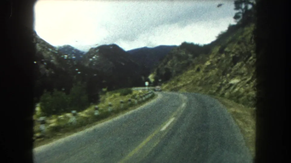 A 1962 Colorado mountain highway curves through green terrain, with guardrail posts, forested slopes, and overcast sky visible ahead.