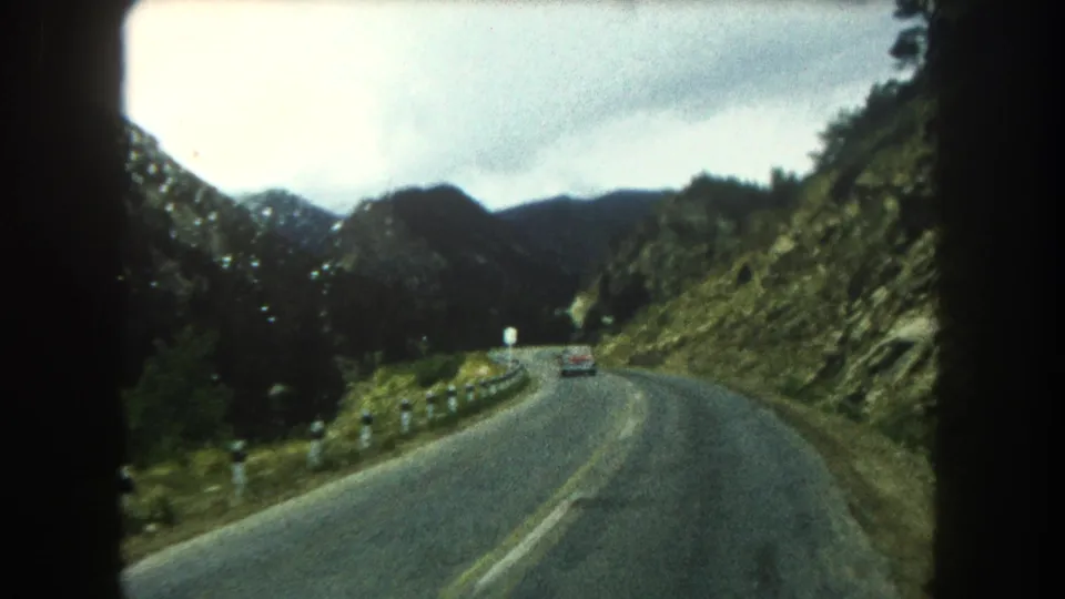 A car travels ahead on a winding 1962 Summit County mountain road, bordered by guardrail posts, green slopes, and distant ridges.