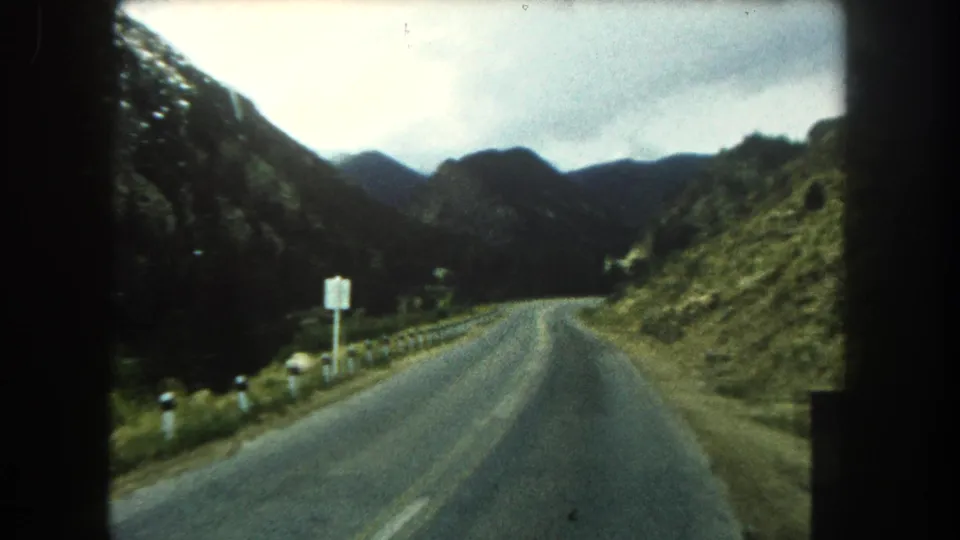 The 1962 Colorado road scene continues along a curving two-lane highway with a roadside sign, guardrail posts, green slopes, and mountains.