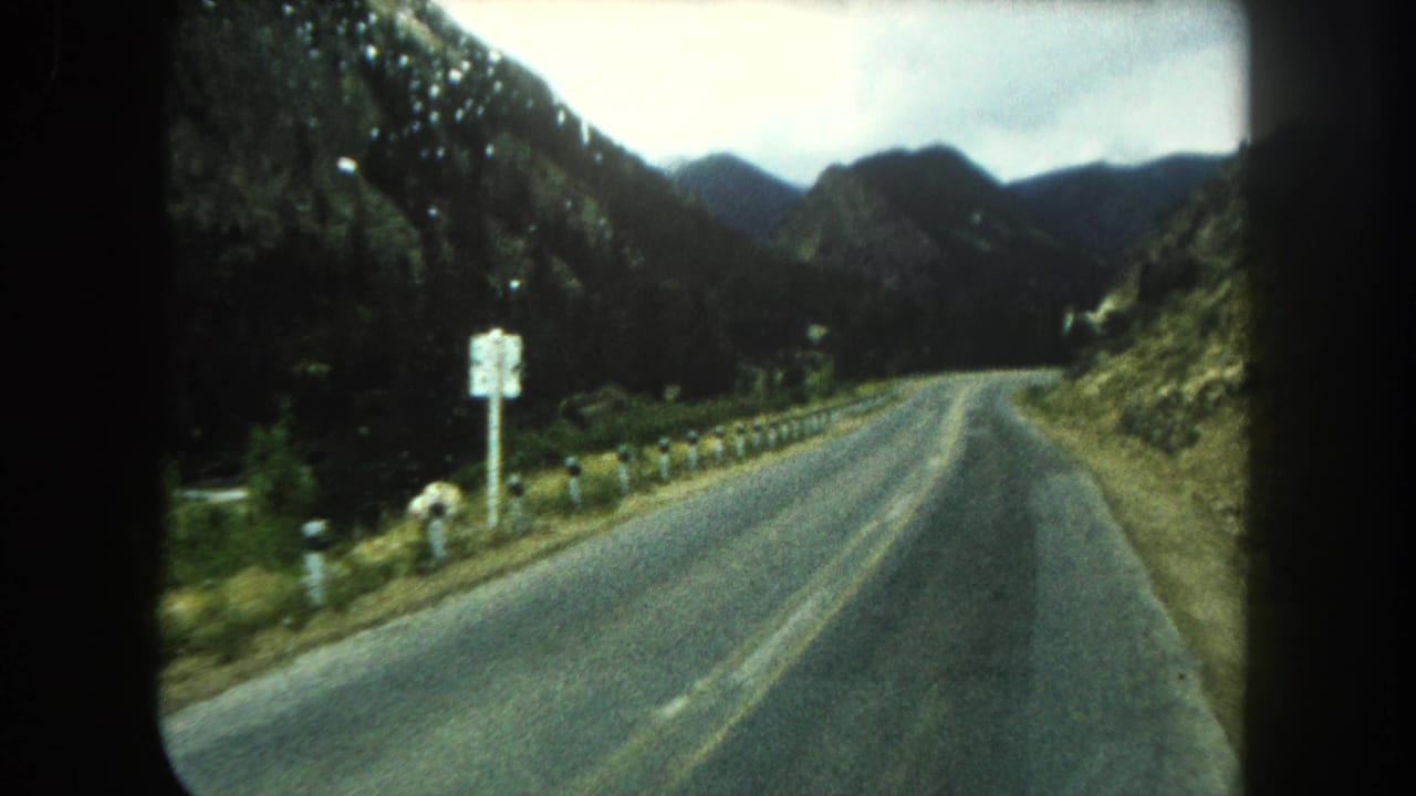 Two-lane mountain road curving through forested slopes under a cloudy sky.